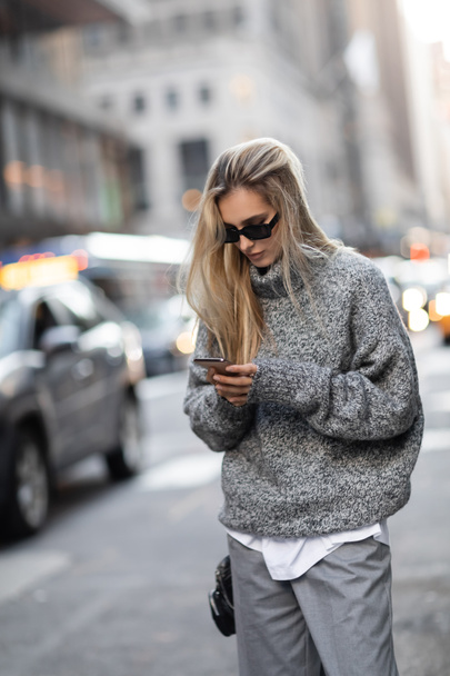 koncentrált nő téli pulóver és napszemüveg segítségével okostelefon a városi utcán New York város  - Fotó, kép