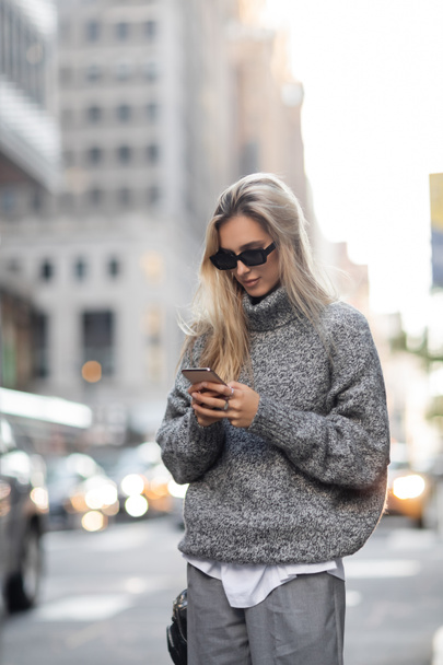блондинка в трикотажном свитере и солнечных очках с помощью смартфона на городской улице в Нью-Йорке  - Фото, изображение