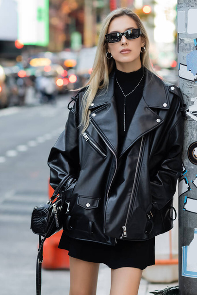 ニューヨークのポケットに手を入れて立つ革のジャケットとサングラスのスタイリッシュな若い女性  - 写真・画像