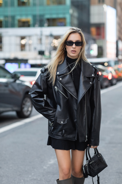 young woman in leather jacket and sunglasses standing with hand in pocket on street of New York  - Fotó, kép