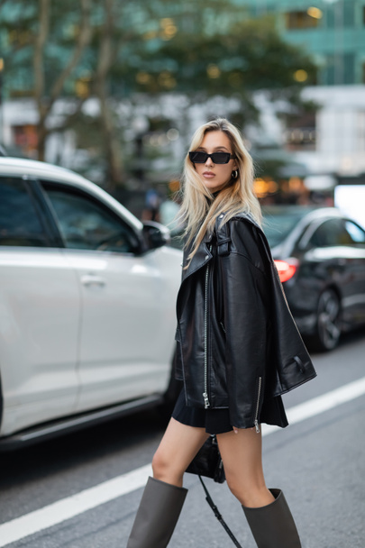 ニューヨークの路上でぼやけた車の近くを歩く革のジャケットと黒のドレスのスタイリッシュな女性  - 写真・画像