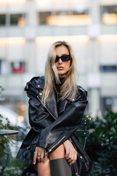 young blonde woman in black leather jacket and sunglasses sitting in outdoor cafe  - Foto, Imagem