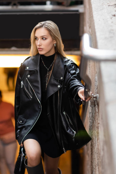 stylish young woman in leather jacket and black dress holding metal handrail near subway entrance in New York  - Photo, image