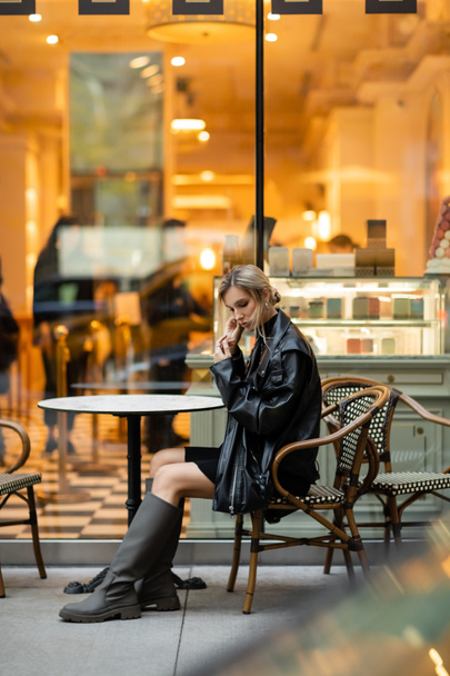 comprimento total de mulher loira em jaqueta de couro preto sentado perto da mesa redonda bistro no café ao ar livre em Nova York  - Foto, Imagem