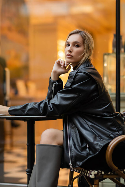 pełna długość młoda kobieta w czarnej skórzanej kurtce siedzi w pobliżu okrągłego stołu bistro w kawiarni na świeżym powietrzu w Nowym Jorku  - Zdjęcie, obraz