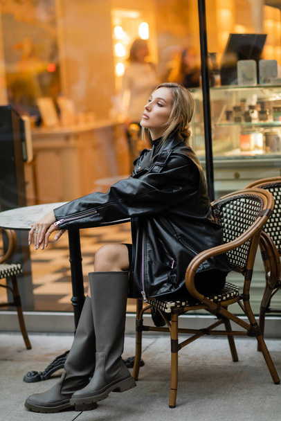 pełna długość stylowej kobiety w czarnej skórzanej kurtce i butach siedzącej w pobliżu stołu bistro w kawiarni na świeżym powietrzu w Nowym Jorku  - Zdjęcie, obraz
