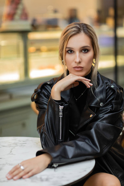 young blonde woman in black leather jacket sitting near bistro table in outdoor cafe in New York  - Photo, Image