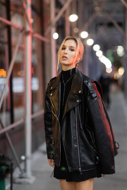 黒の革のジャケットを着たブロンドの女性が夜のニューヨークの通りに立っている - 写真・画像