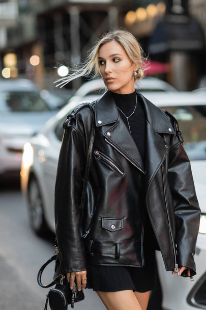 stylowa kobieta w skórzanej kurtce i czarnej sukience spacerująca w pobliżu zamazanych samochodów na ulicy w Nowym Jorku - Zdjęcie, obraz