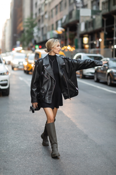 longitud completa de la mujer rubia en chaqueta de cuero negro y vestido de captura de taxi en la calle en Nueva York  - Foto, imagen