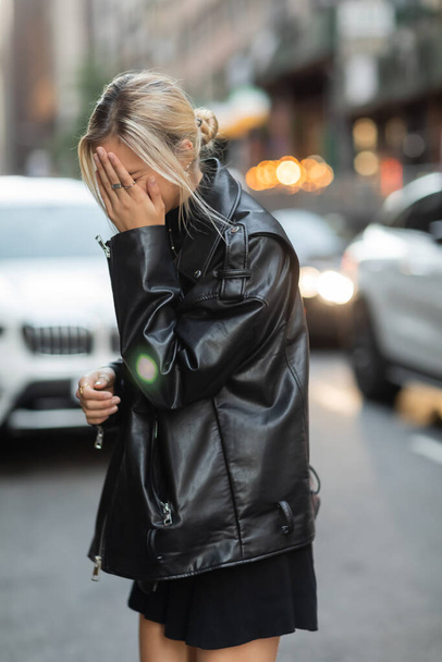 блондинка в черной кожаной куртке покрывает лицо рукой на улице в Нью-Йорке  - Фото, изображение
