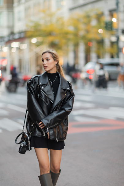 elegante donna in giacca di pelle e vestito a piedi vicino a auto sfocate in strada nella città di New York  - Foto, immagini