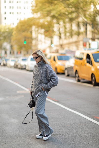 longitud completa de mujer joven bonita en traje de invierno caminando con bolso de moda cerca de coches borrosos en Nueva York - Foto, Imagen