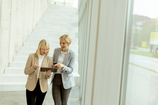 Дві ділові жінки ходять з ноутбуком і мобільним телефоном в офісному коридорі
 - Фото, зображення