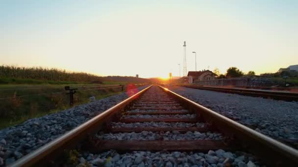 Tren yolu boyunca, köy tren istasyonunu geçerek güneşe doğru ilerliyoruz. Altın ışıkta kırsal demiryolu ve tren istasyonu. Tren raylarının ve rayların detaylı görüntüsü. - Video, Çekim