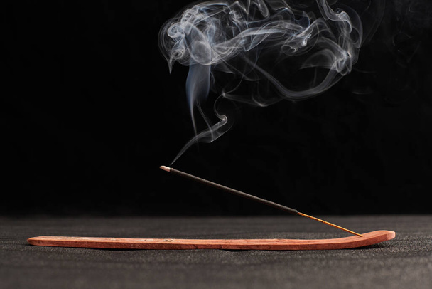 Smoke curls of incense stick in wooden holder for relaxation and meditation exercises, black background. Aromatherapy session with burning aroma stick, pleasant aroma and incense - Foto, Imagem