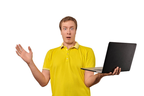 Έκπληκτος τύπος κρατώντας φορητό υπολογιστή στο χέρι απομονώνονται σε λευκό φόντο. Αστείος σοκαρισμένος νεαρός άνδρας με κίτρινο μπλουζάκι κρατώντας σημειωματάριο στο χέρι. Αστείος τύπος με μπερδεμένο βλέμμα λόγω πωλήσεων και εκπτώσεων - Φωτογραφία, εικόνα