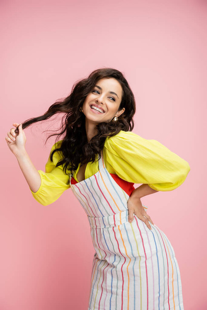 coquettish housewife in bright yellow blouse and apron holding wavy hair and posing with hand on hip isolated on pink - Фото, изображение