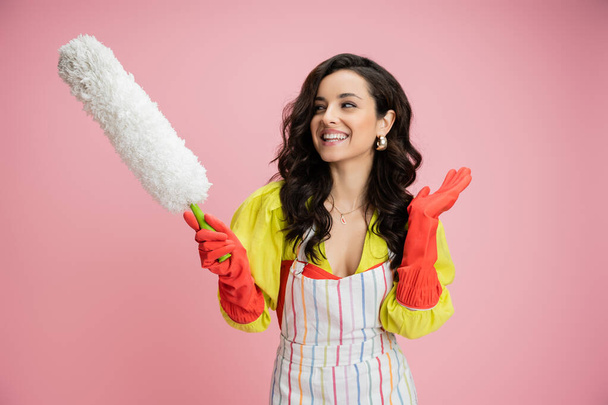 улыбающаяся домохозяйка в полосатом фартуке и красных резиновых перчатках держа белую пыль кистью и размахивая рукой изолированы на розовый - Фото, изображение