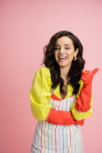 excited brunette housewife in red rubber gloves laughing and pointing with thumb isolated on pink - Foto, Imagen