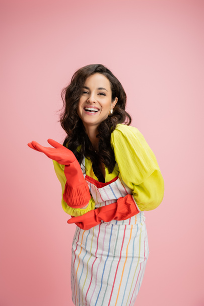 excitée brune femme au foyer en gants de caoutchouc rouge riant et pointant avec la main isolée sur rose - Photo, image