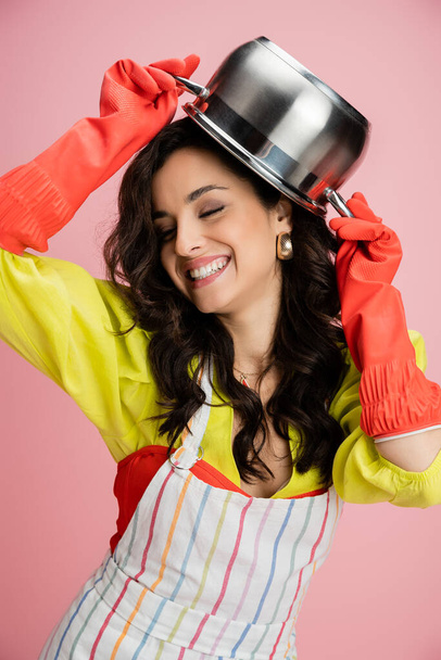 cheerful woman in red rubber gloves having fun while holding saucepan above head isolated on pink - Zdjęcie, obraz