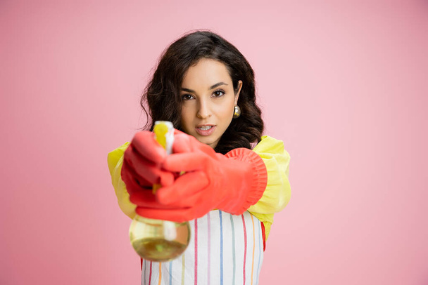 young brunette housewife in red rubber gloves holding spray bottle and looking at camera isolated on pink - Photo, Image