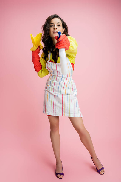 longitud completa de la mujer en delantal rayado y guantes de goma sosteniendo botella de spray y trapo mientras mira a la cámara en rosa  - Foto, Imagen