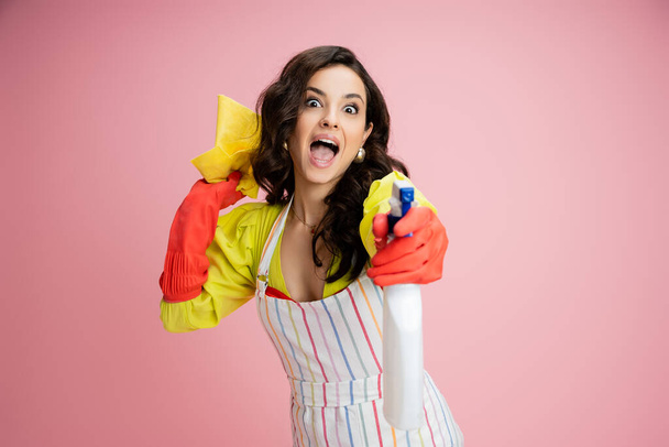 excited housewife having fun while holding rag and spraying from bottle at camera isolated on pink - Fotoğraf, Görsel
