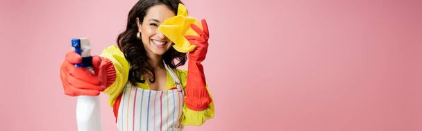 cheerful housewife in red rubber gloves obscuring face with rag and spraying from bottle isolated on pink, banner - Photo, image