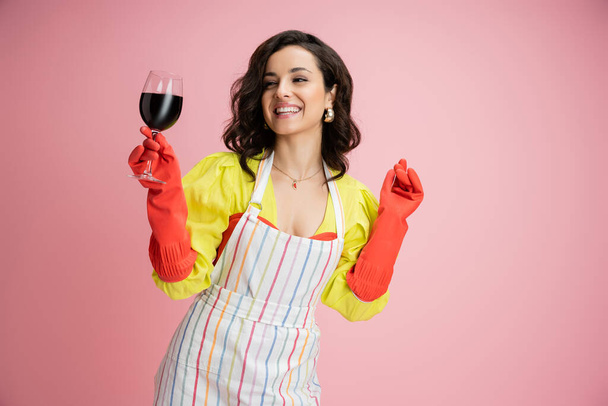 kokette huisvrouw in rood rubber handschoenen en schort glimlachen en houden glas wijn geïsoleerd op roze - Foto, afbeelding