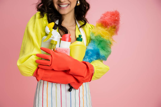vista recortada de ama de casa sonriente en guantes de goma roja sosteniendo un montón de artículos de limpieza aislados en rosa - Foto, Imagen