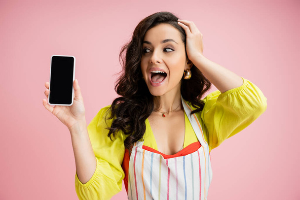mujer asombrada en blusa amarilla y delantal rayado tocando la cabeza mientras sostiene el teléfono celular con pantalla en blanco aislado en rosa - Foto, imagen