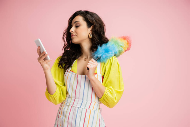 displeased housewife in striped apron holding colorful dust brush and looking at mobile phone isolated on pink - Photo, Image