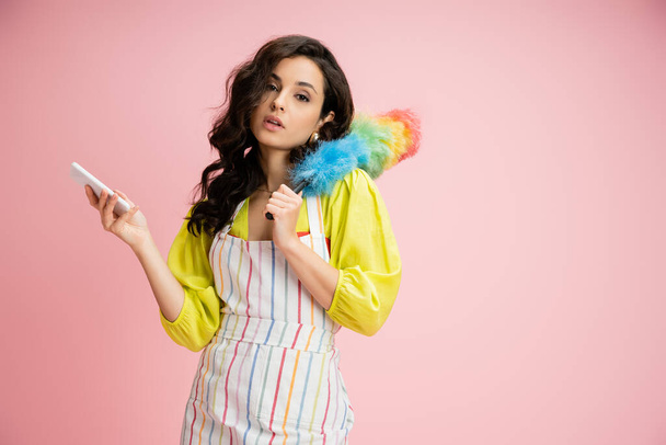 pretty brunette housewife in striped apron holding smartphone and colorful dust brush while looking at camera isolated on pink - Photo, Image