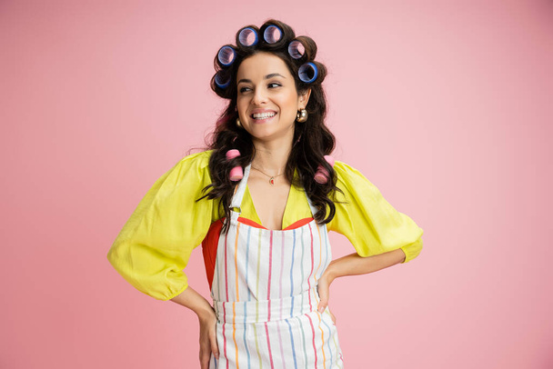 cheerful housewife in yellow blouse and hair curlers looking away while holding hands on hips isolated on pink - Photo, Image