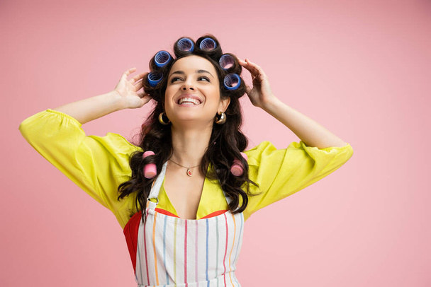 cheerful young woman in yellow blouse and apron touching hair curlers and looking away isolated on pink - Photo, Image
