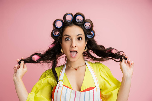 surprised housewife in hair curlers and yellow blouse holding hair and looking at camera isolated on pink - Photo, Image