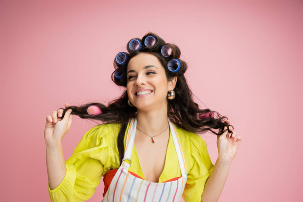flirty housewife in striped apron and hair curlers pulling hair and smiling while looking away isolated on pink - Фото, зображення