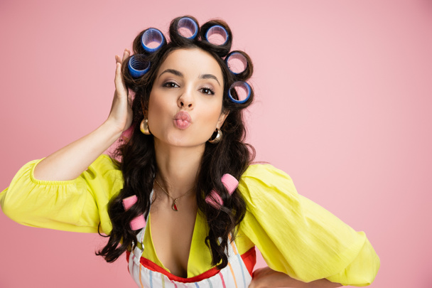 young housewife in yellow blouse touching hair curlers and pouting lips isolated on pink - Photo, Image