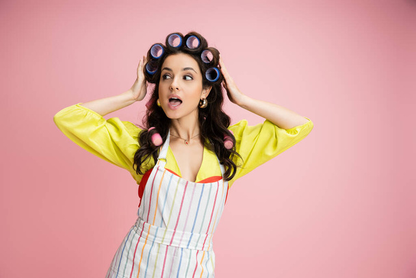 изумленная и любопытная домохозяйка в полосатом фартуке регулирует волосы бигуди и смотрит в сторону изолированы на розовый - Фото, изображение