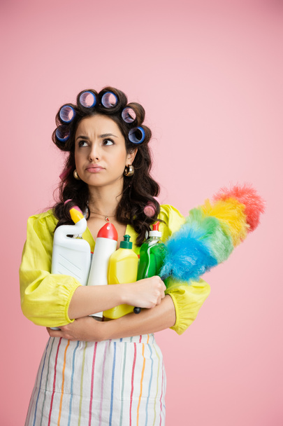 displeased housewife with plenty of cleaning supplies and colorful feather duster looking away isolated on pink - Photo, Image