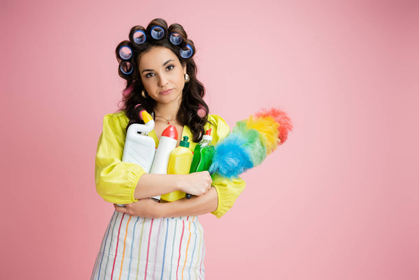 bored housewife with hair curlers and different cleaning supplies looking at camera isolated on pink - Photo, Image