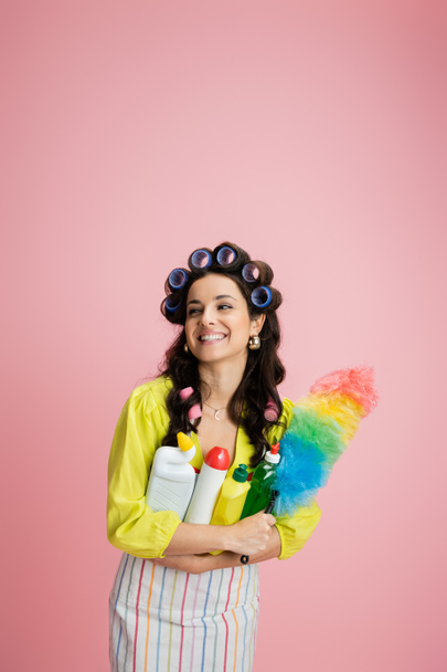 pretty housewife with hair curlers and cleaning supplies smiling and looking away isolated on pink - Photo, Image