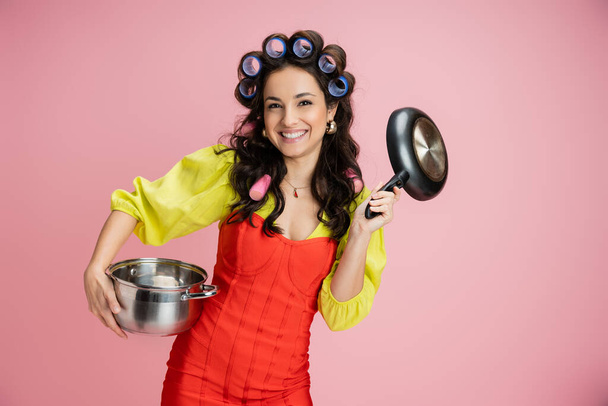 веселая домохозяйка в красном корсетном платье и бигуди для волос держа кастрюлю и сковородку, глядя на камеру, изолированную на розовый - Фото, изображение