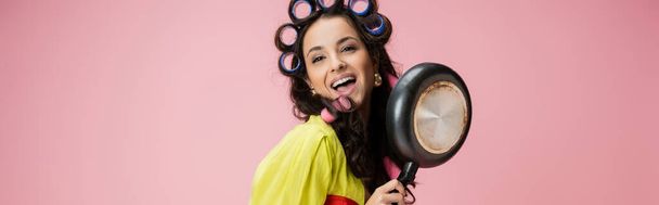 happy housewife in hair curlers looking at camera near frying pan isolated on pink, banner - Foto, Imagen