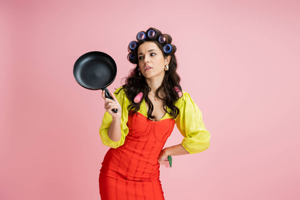 bored housewife in hair curlers and red corset dress looking at frying pan isolated on pink - Fotó, kép