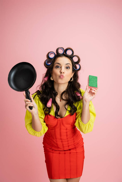 young housewife in hair curlers pouting lips while holding frying pan and sponge isolated on pink - Photo, Image