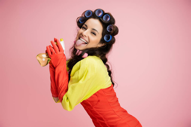 excited and pretty housewife in hair curlers and red corset dress holding spray bottle and sticking out tongue isolated on pink - Photo, Image