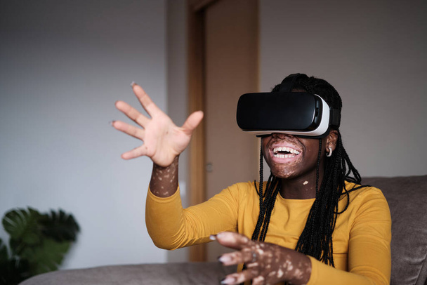 陽気なアフリカ系アメリカ人女性でVRゴーグルは仮想現実と相互作用しながら、自宅でソファの上に休んで楽しい - 写真・画像